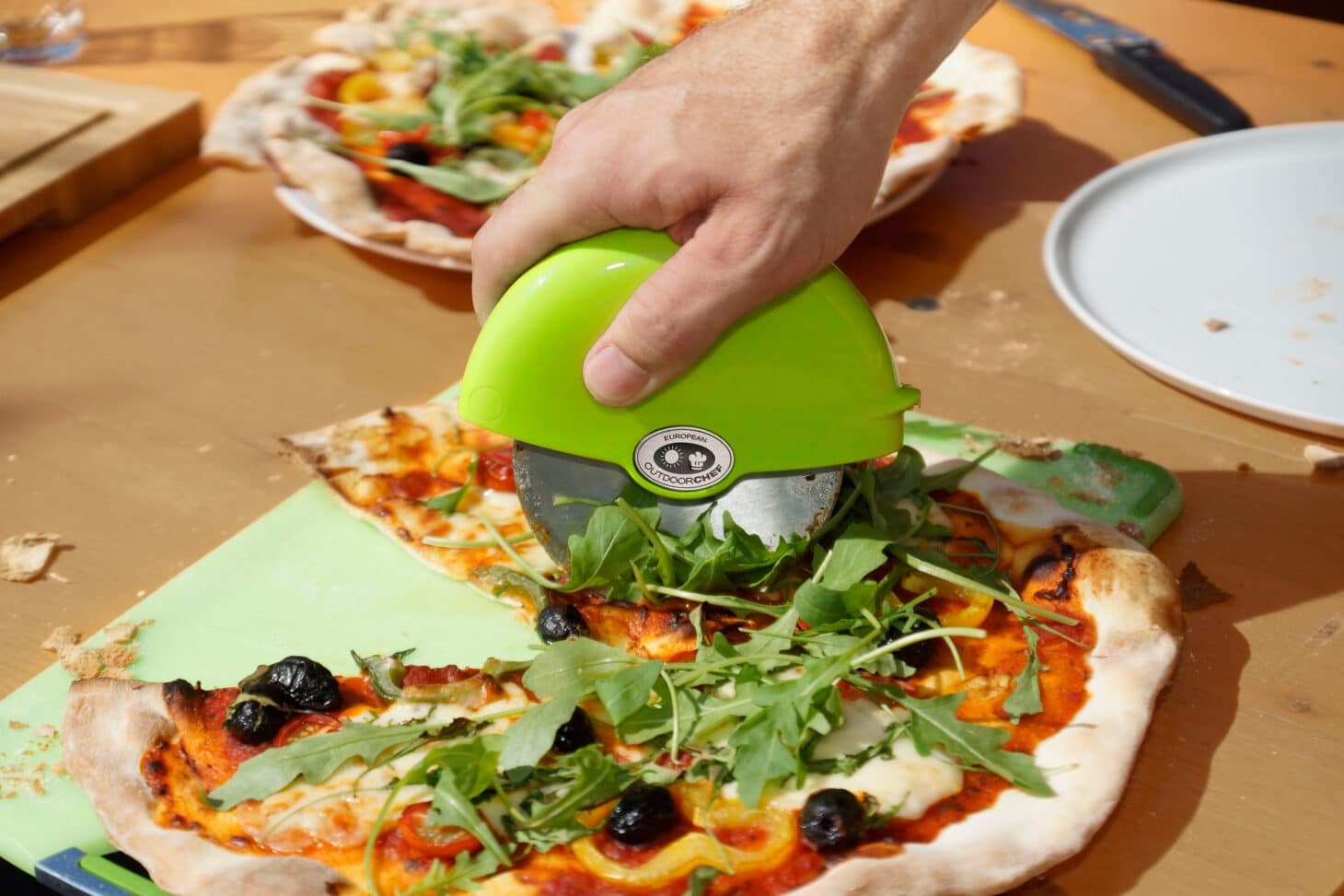 Outdoorchef Pizzaschneider schneidet frisch gebackene Pizza mit Rucola, Oliven und Gemüse