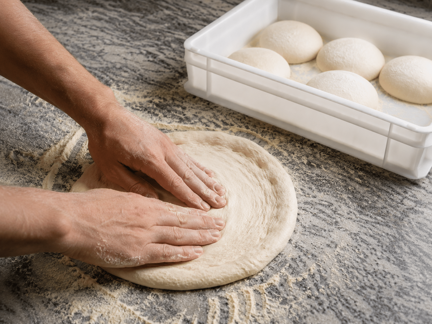 Eine Person formt einen runden Pizzateig mit den Händen von innen nach außen auf einer bemehlten Arbeitsplatte; im Hintergrund steht eine weiße Teigbox mit fünf Teiglingen